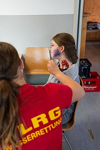Einsatzkraft der DLRG beim Schminken einer weiteren Person mit einem blauen Fleck und einer größeren Verletzung am Hals