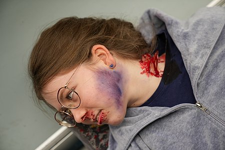 Person mit mehreren dargestellten Verletzungen am Kopf und Hals am Boden liegend dargestellt